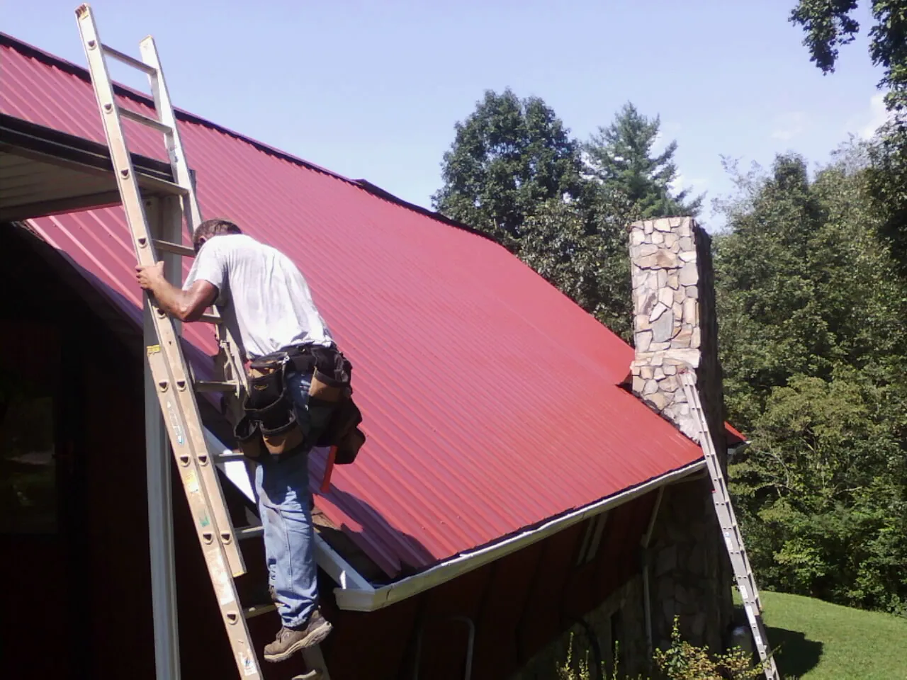 Roofer on a ladder beside a red metal roof and stone chimney — Soffit & Fascia Repair by Roofing Brothers in Neptune